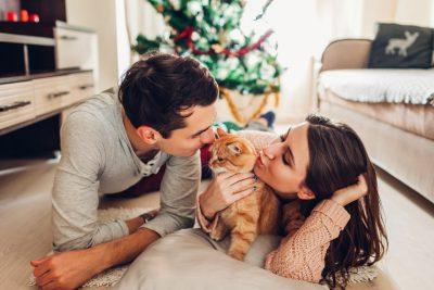 A man and woman kissing their cat on the floor of a living room in front of their Christmas tree